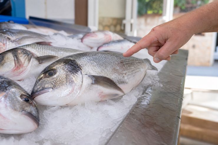 Dorada fresca sobre hielo mientras una persona señala la pieza en el mostrador